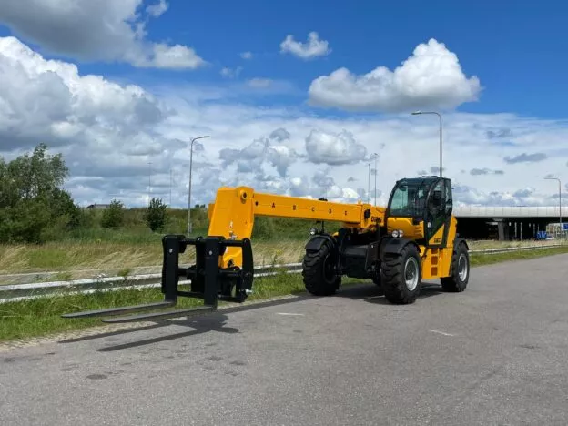 Yellow telehandler with extended forks on a road against a backdrop of trees and a bright blue sky with clouds.