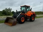 Bright orange Doosan DL200 front loader on a road with a green, tree-lined background under a cloudy sky.