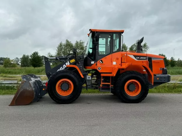 Bright orange Doosan DL200 wheel loader parked on a road with a cloudy sky backdrop.