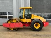 Yellow and red construction road roller parked in front of a grey industrial building with large windows.