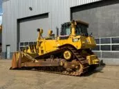 Yellow Caterpillar (CAT) D8T bulldozer parked in front of a large industrial garage door at a construction site.