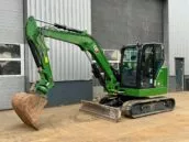 Green excavator parked outside a grey industrial building with large windows and a metal facade.