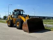 Yellow Caterpillar wheel loader parked on a road near industrial buildings under a clear blue sky.