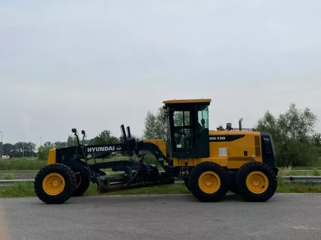 Yellow Hyundai HG130 motor grader parked on a road against a backdrop of trees and overcast sky.