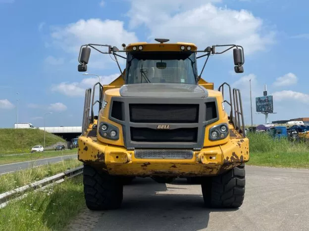 Front view of a Bell construction dump truck on the roadside with a highway and billboard in the background on a sunny day.