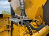 Close-up of yellow construction machinery with hydraulic hoses and safety warnings against a blue sky background.